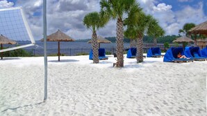 Een strand met een volleybalnet, palmbomen en blauwe ligstoelen.