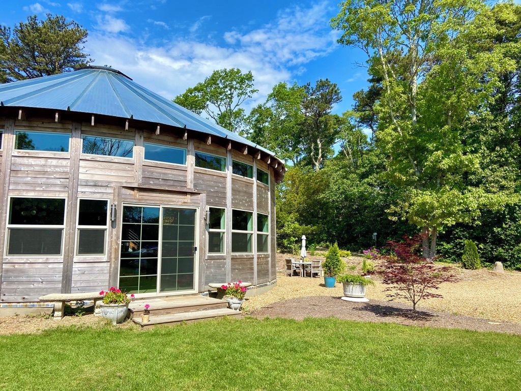 A wooden building with a curved roof and large windows, surrounded by a well-maintained garden with various plants and a patio area.