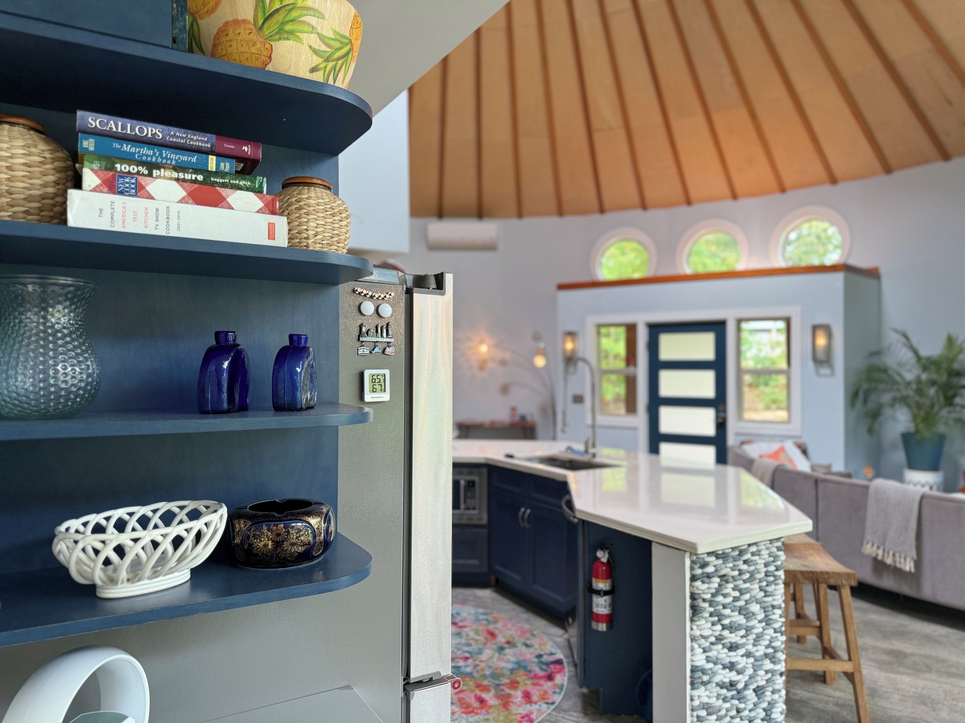 A modern kitchen with a built-in refrigerator, shelves with books and decorative items, a round island with a sink, and a wooden ceiling.