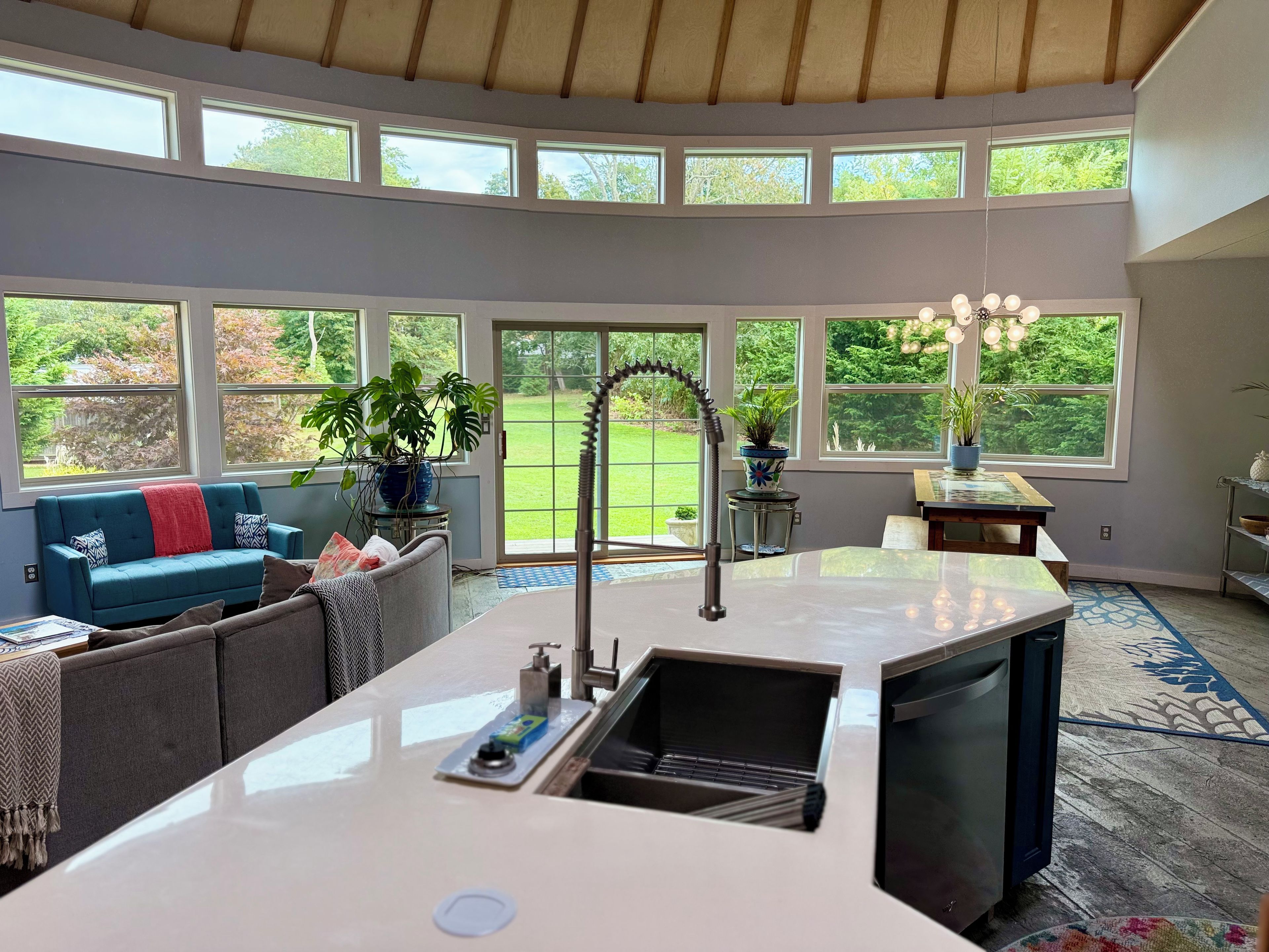 A modern kitchen with a large island, a dining area with a table and chairs, and a view of a green garden through large windows.