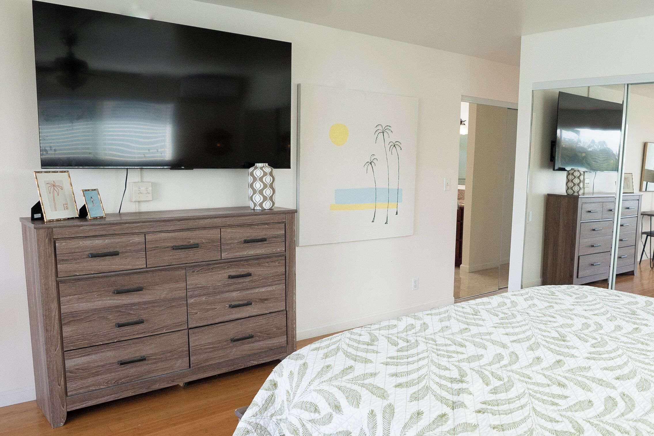 Un dormitorio con cama, cómoda, televisor montado en la pared y un cuadro.