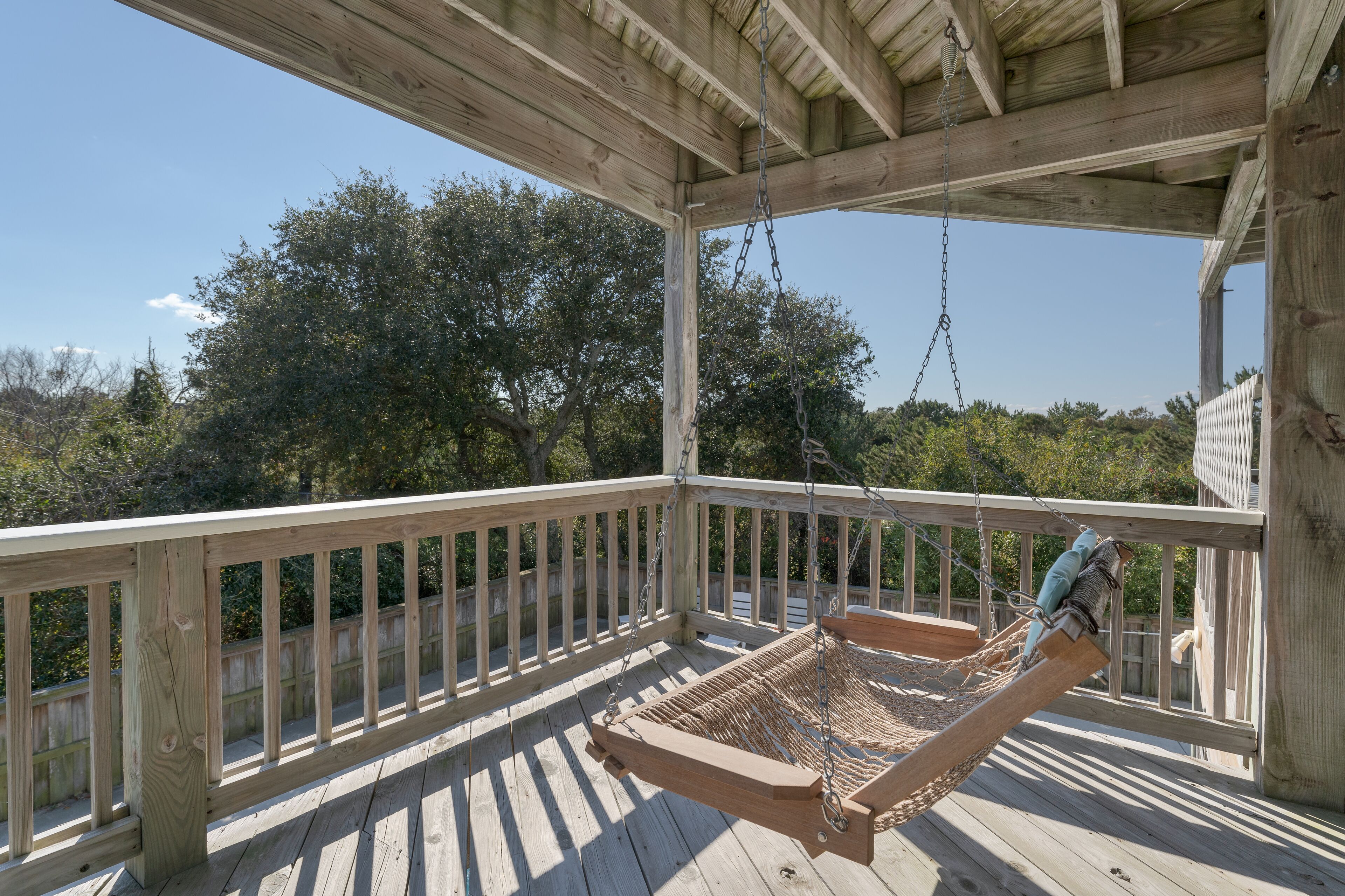 A wooden deck with a hammock swing, a wooden railing, and a view of trees and sky.