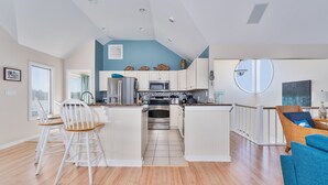 A modern kitchen with white cabinets, a central island, and a dining area with a wooden chair. - 1623 - Breakaway - Bring the Pup! Corolla Escape w/ Pool & Hot Tub (Corolla)