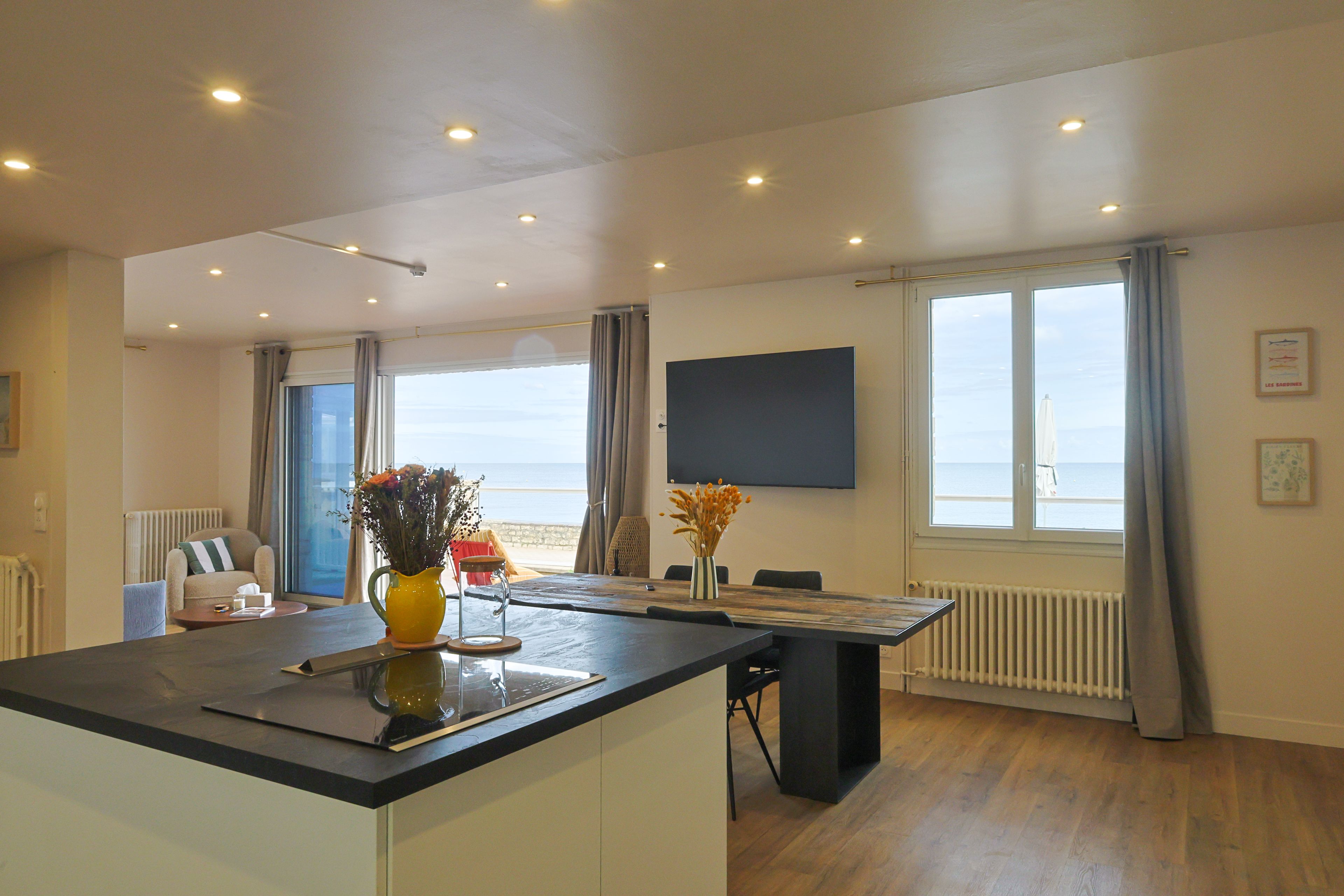 Une cuisine moderne dotée d’un îlot central, d’un coin repas avec une table et des chaises, d’une vue sur la mer grâce à de grandes fenêtres et d’un téléviseur à écran plat fixé au mur.