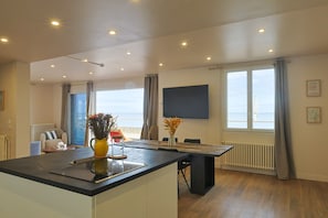 A modern kitchen with a central island, a dining area with a table and chairs, a view of the sea through large windows, and a flat-screen TV mounted on the wall. - Le Clos Normand (Saint-Aubin-sur-Mer)