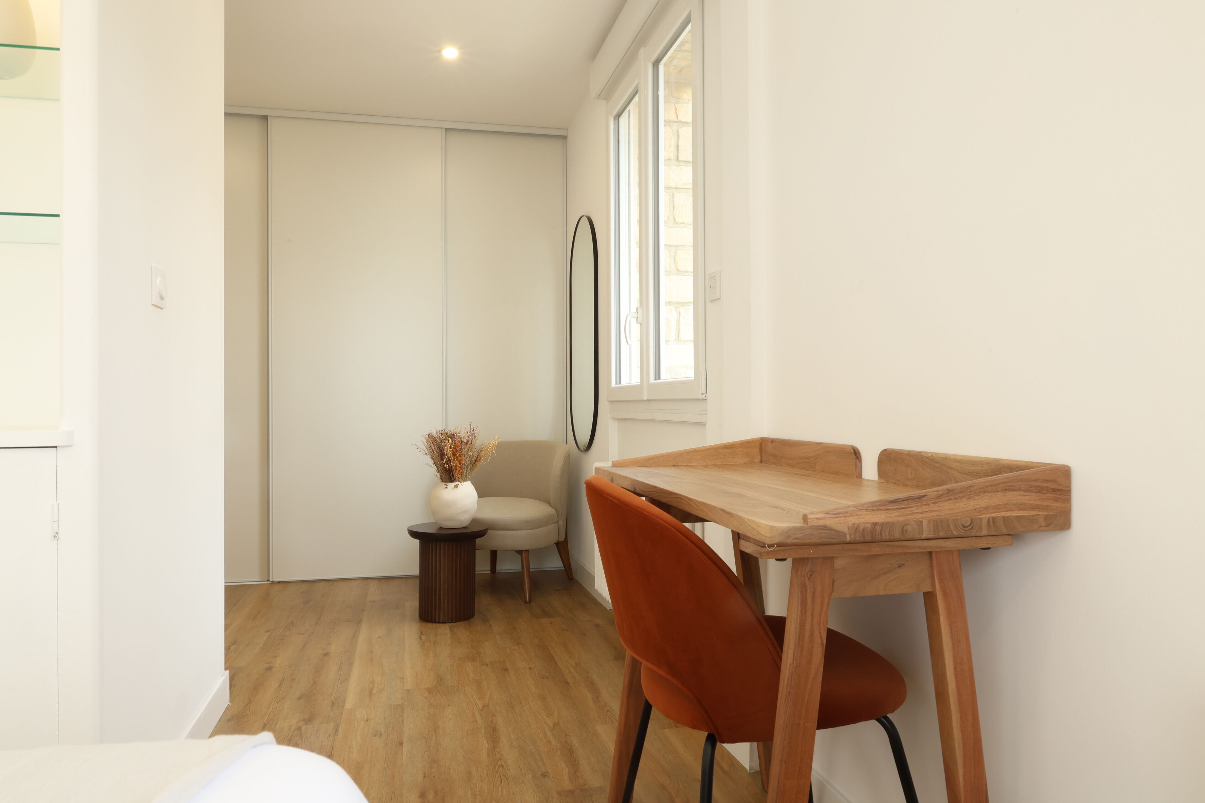 Une pièce comprenant une table à manger en bois, une seule chaise orange, une petite table d’appoint avec un vase, et une grande fenêtre avec des stores.