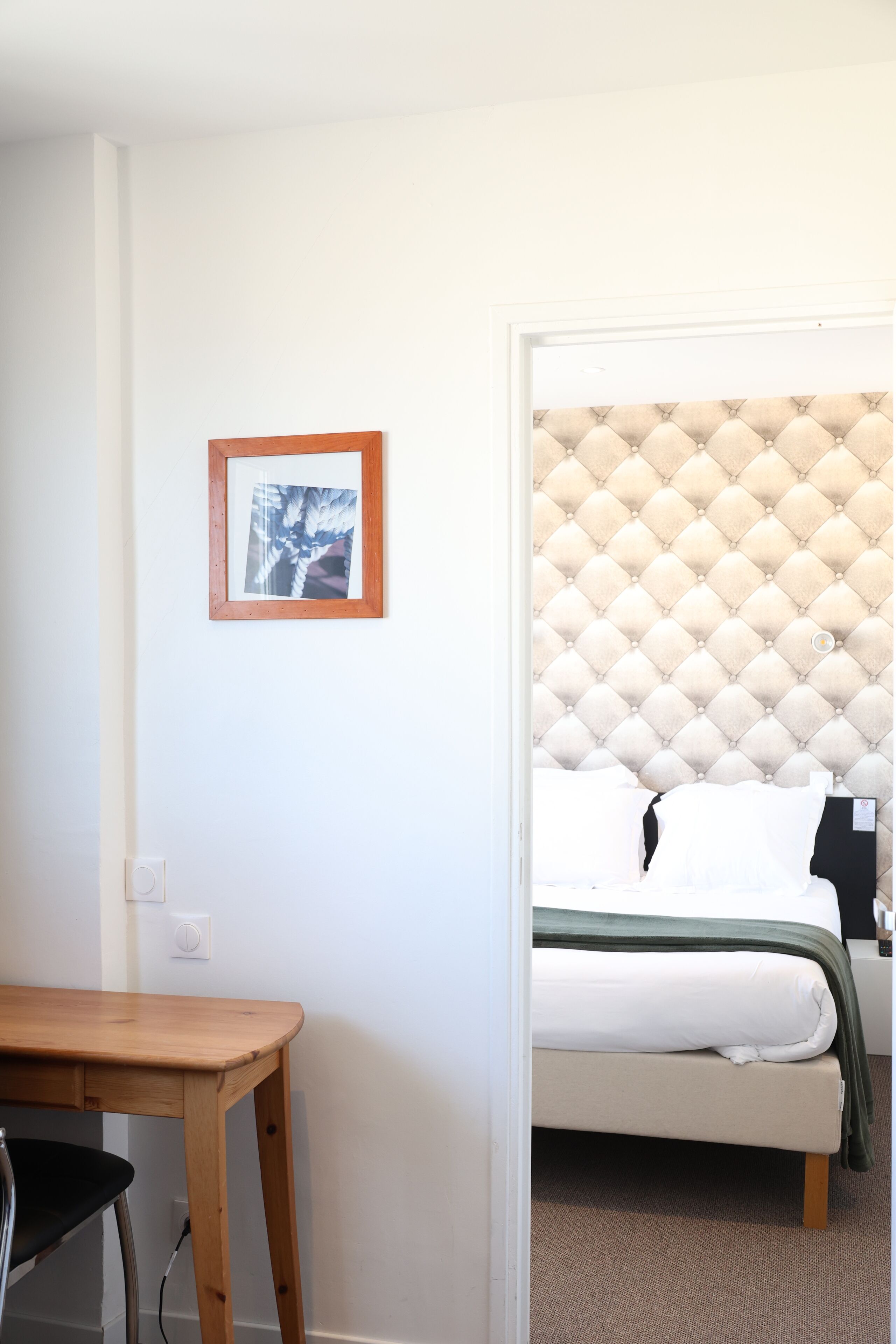 A bedroom with a tufted headboard, a bed with white bedding, a wooden desk, and a framed picture on the wall.