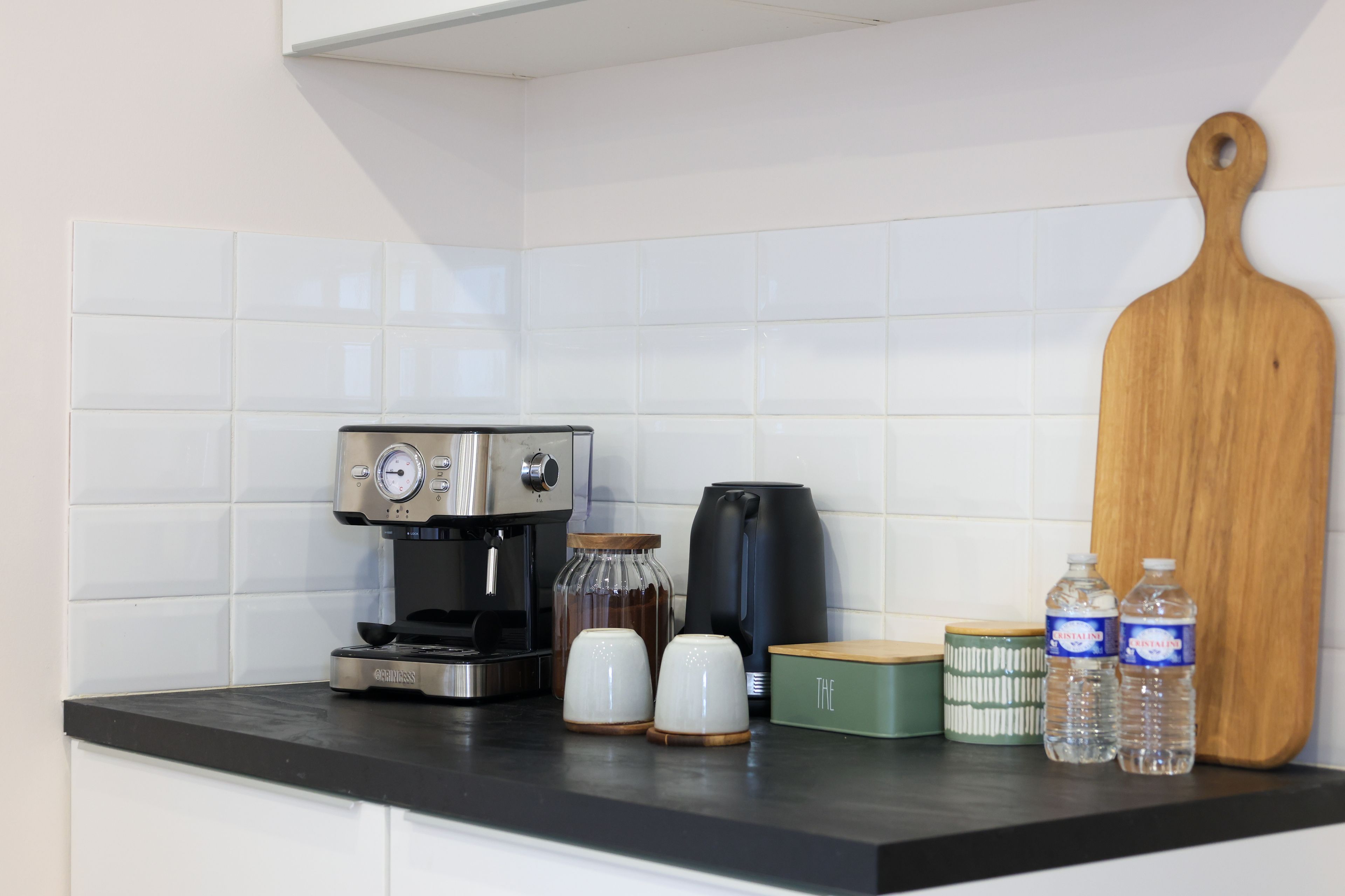 Un comptoir de cuisine avec une machine à café, deux tasses blanches, une bouilloire électrique noire, une planche à découper en bois et deux bouteilles d’eau.