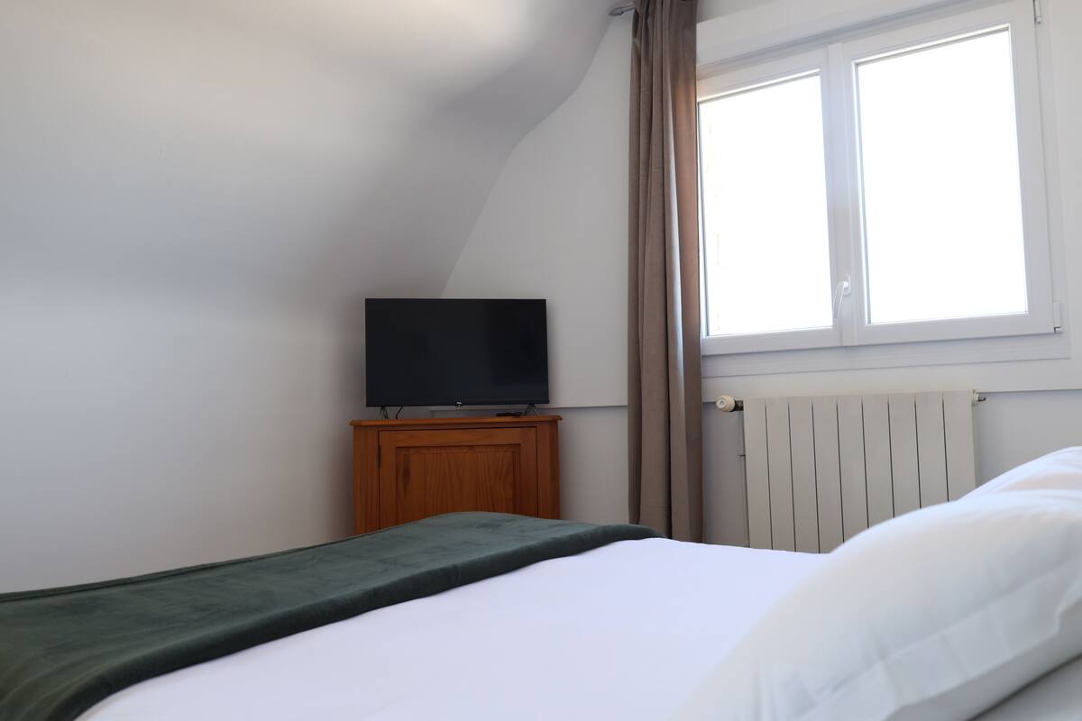 A bedroom with a bed, a television on a wooden stand, a window with curtains, and a radiator.
