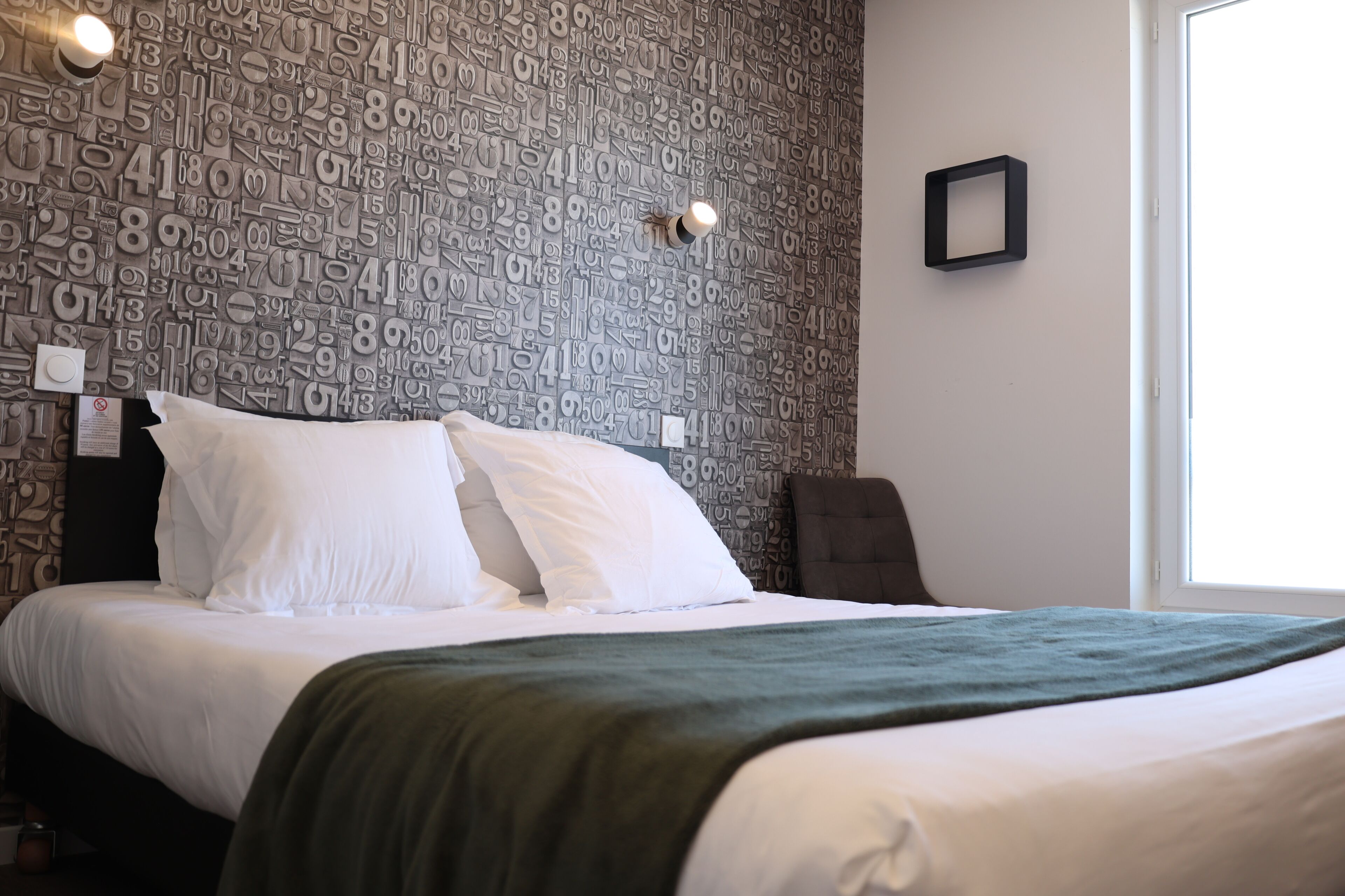 A neatly made bed with white linens and a green throw blanket, a wall with a numerical pattern, and a single framed picture on the wall.