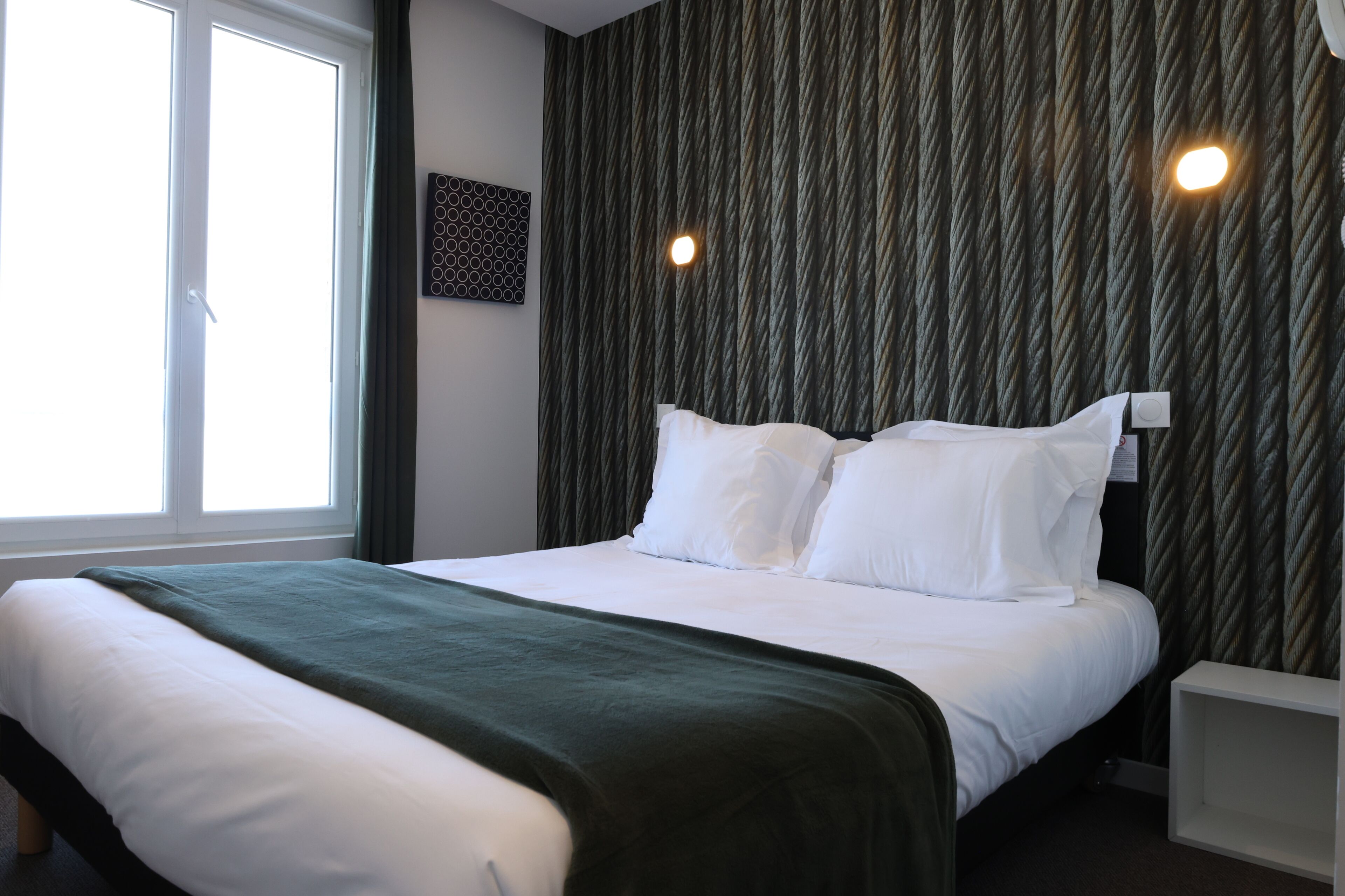 Een net opgemaakt bed met witte lakens en een groene deken, een raam met gordijnen, en een muur met een structuur.