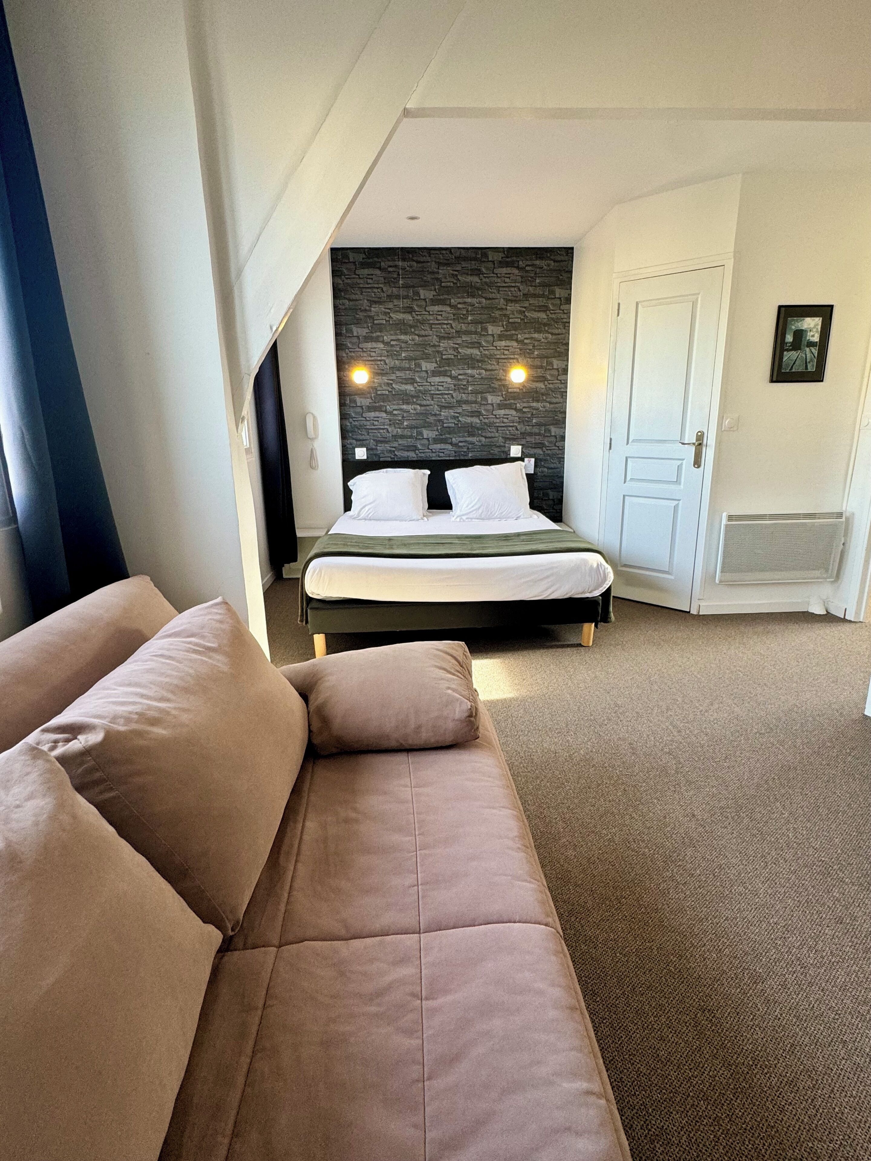 A modern hotel room with a large bed, a built-in headboard, a dark stone accent wall, a white door, and a beige carpet.