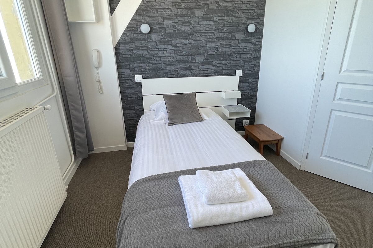 A neatly arranged hotel room with a bed, bedside tables, a nightstand, a radiator, a door, and a stone-patterned wall.