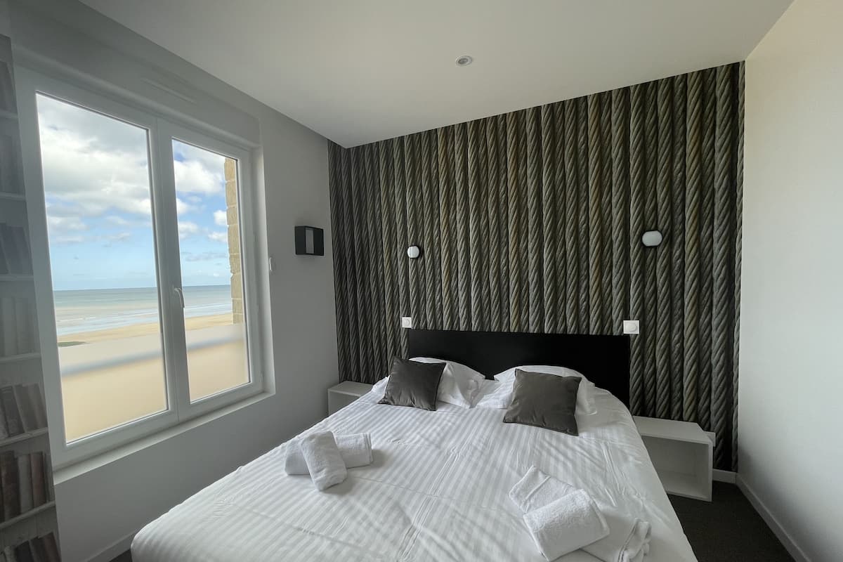 A hotel room with a large window, a bed with white linens, a headboard, and a textured wall.