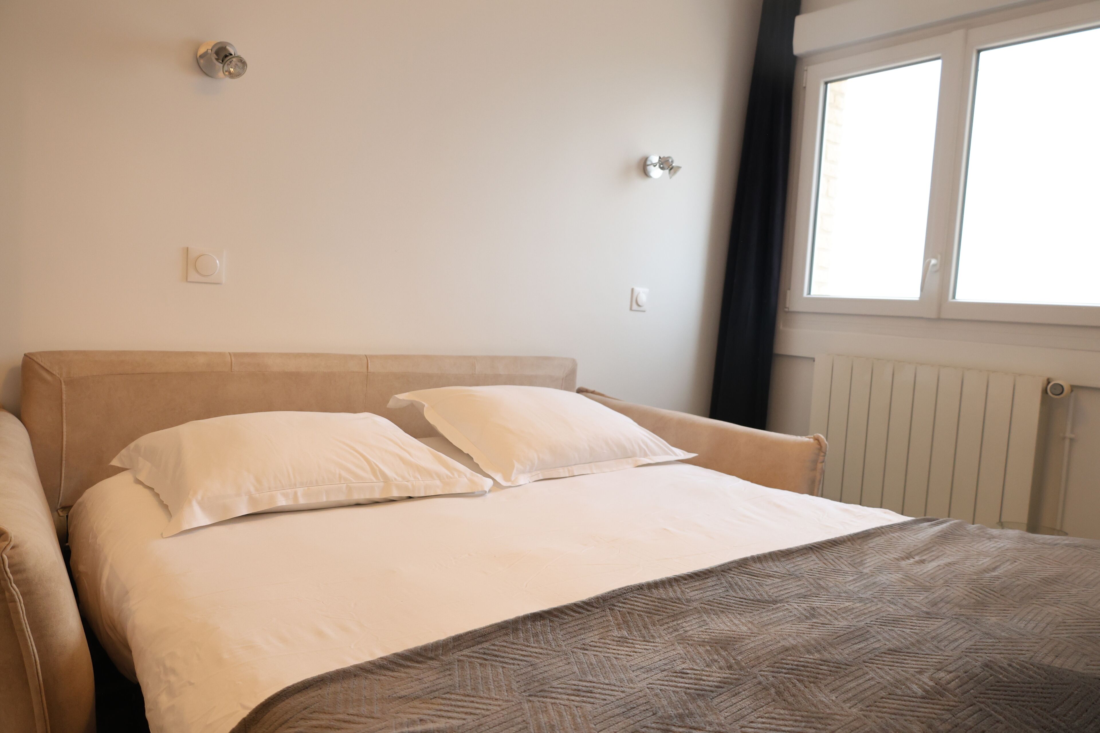 A bedroom with a bed, two pillows, a headboard, a window, and a radiator.