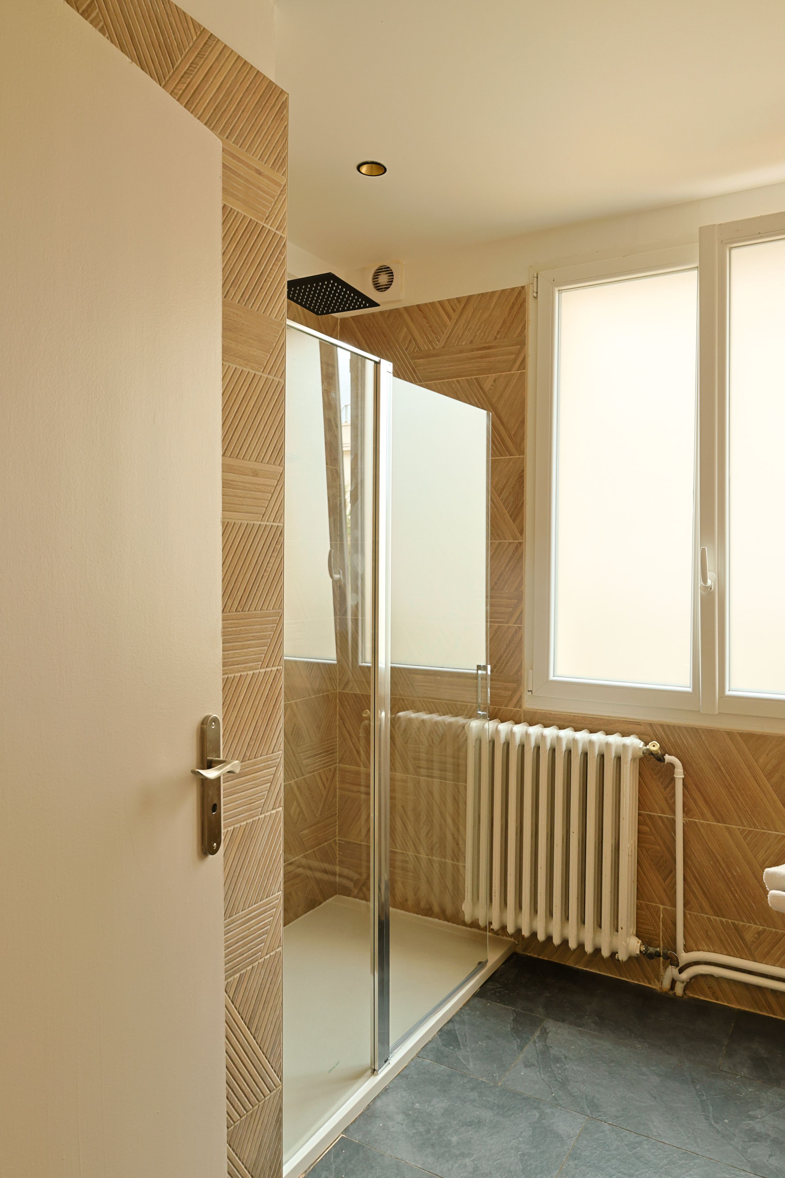 Casa de banho com cabine de duche em vidro, radiador branco e janela com persianas.