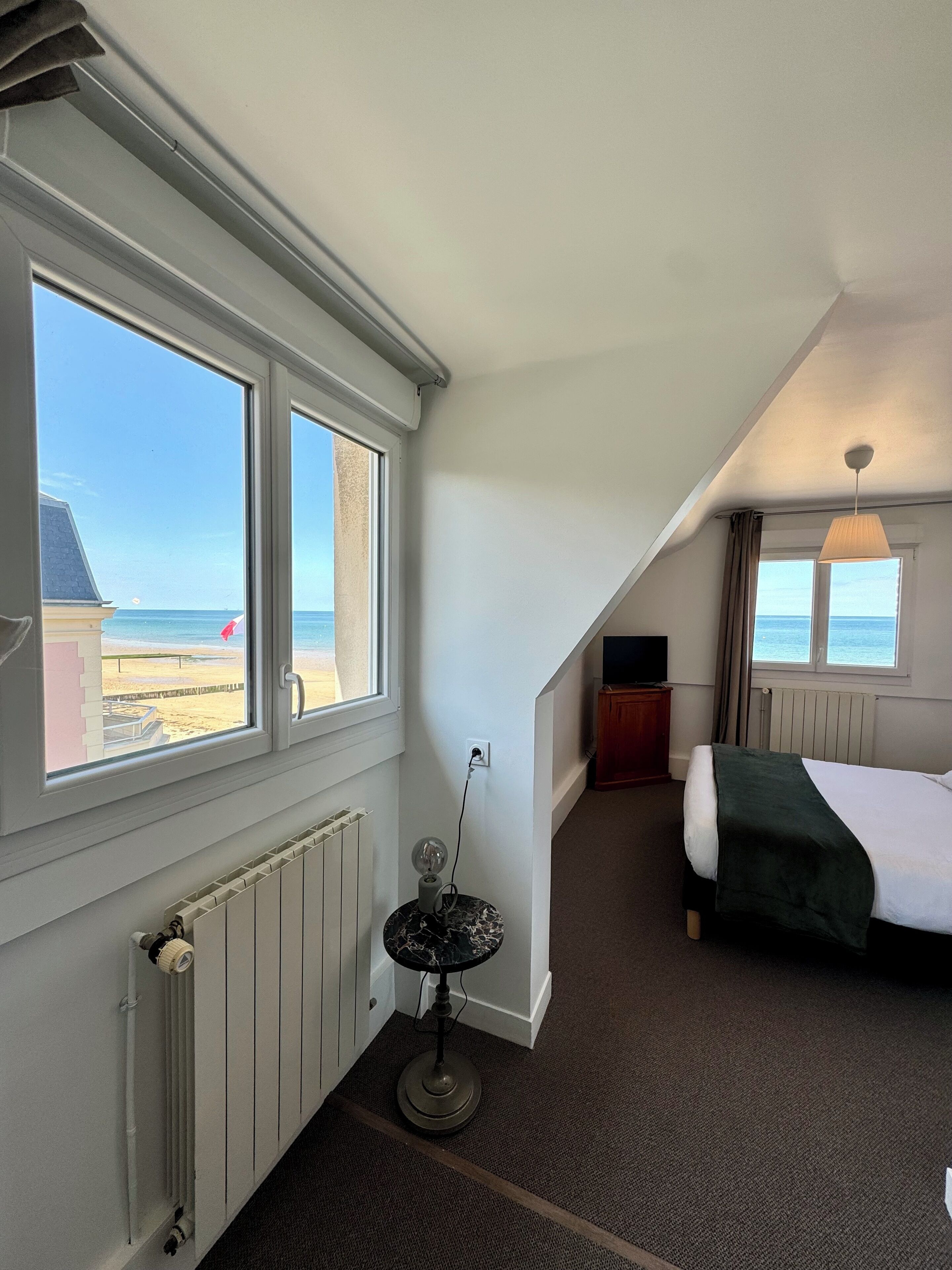 Habitación de hotel con una cama, una mesita con lámpara, un radiador y vistas a la playa.
