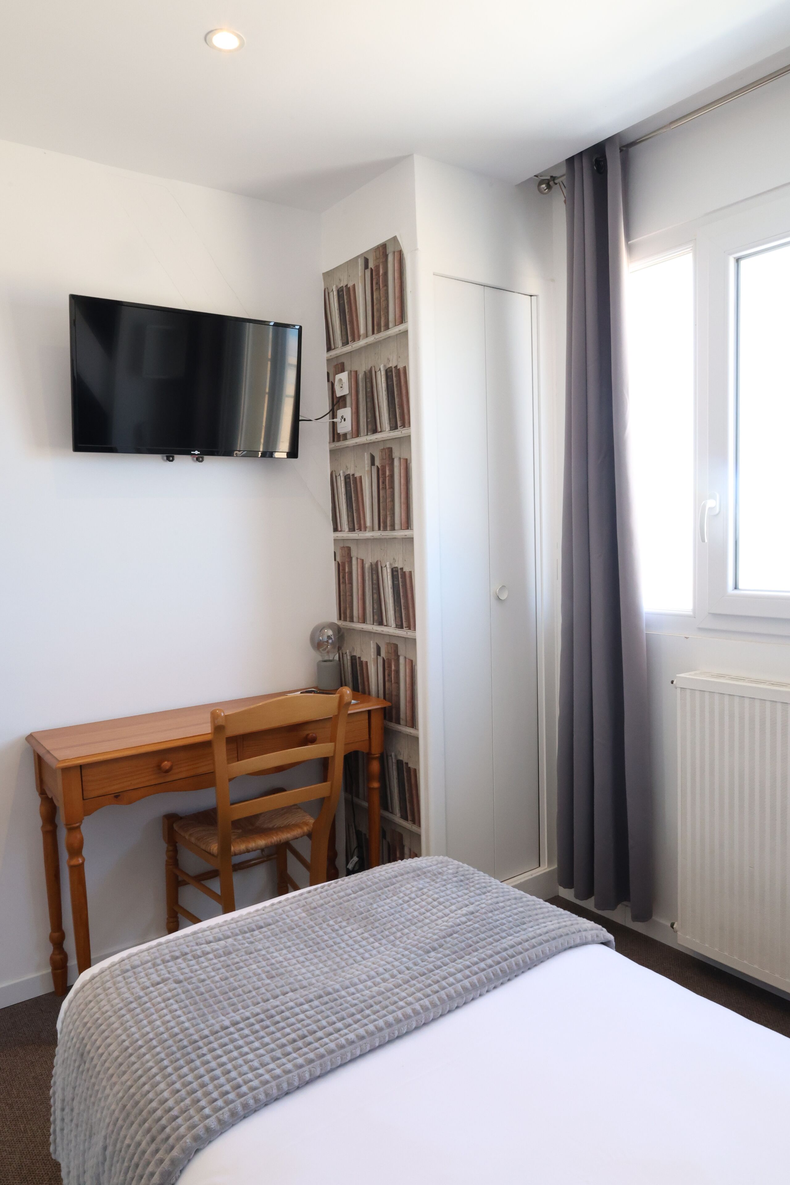 Quarto com cama, escrivaninha, cadeira, estante de livros, televisão e janela com cortinas.