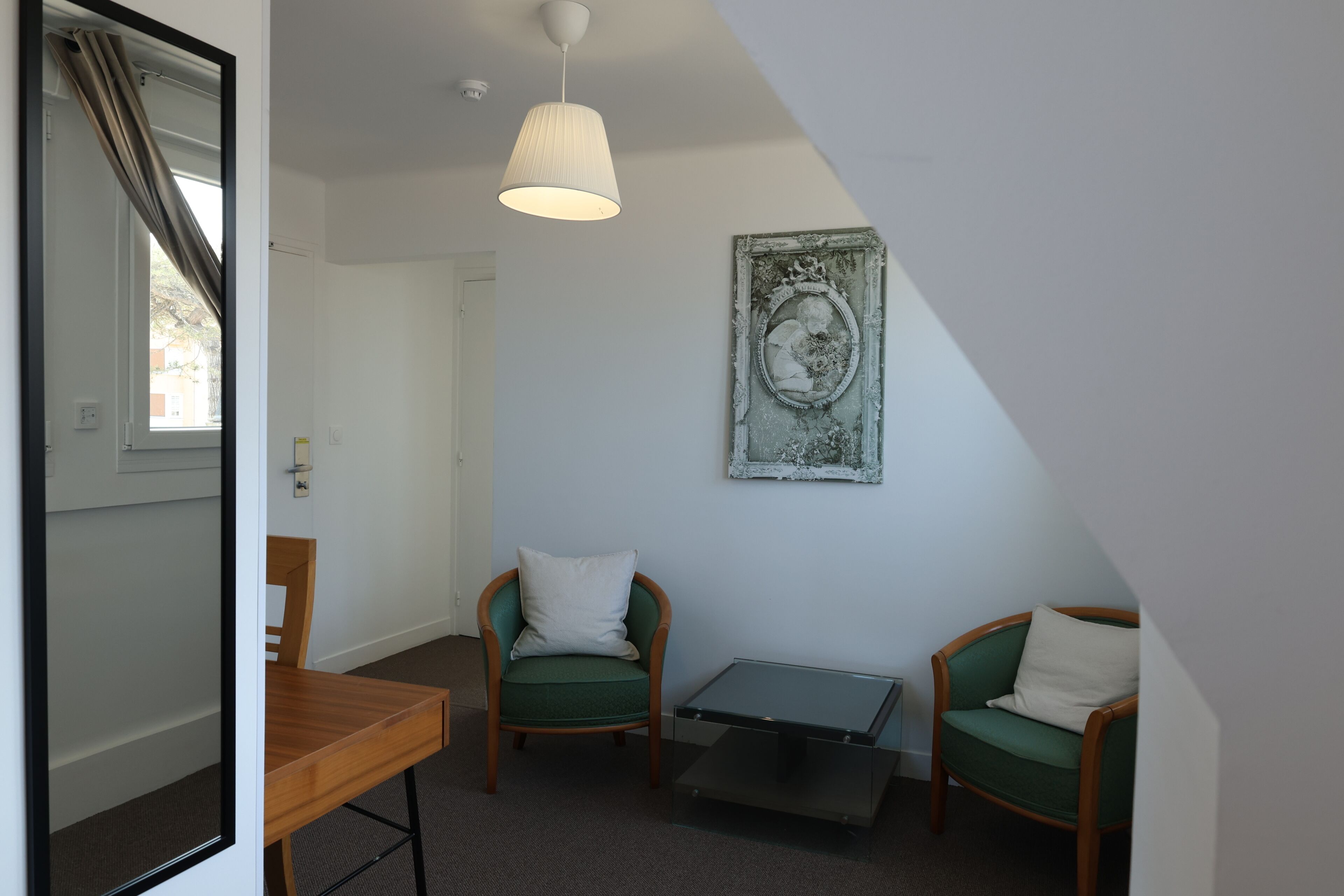 A room with a wooden table, two green armchairs, a small glass table, a white lamp, and a framed picture on the wall.