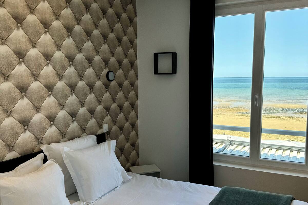 A hotel room with a tufted headboard, a bed with white linens, a green throw blanket, and a view of the beach through a large window.