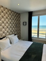 Habitación de hotel con tablero de cabecera capitonado, cama con ropa blanca, manta verde y vista a la playa a través de un gran ventanal.