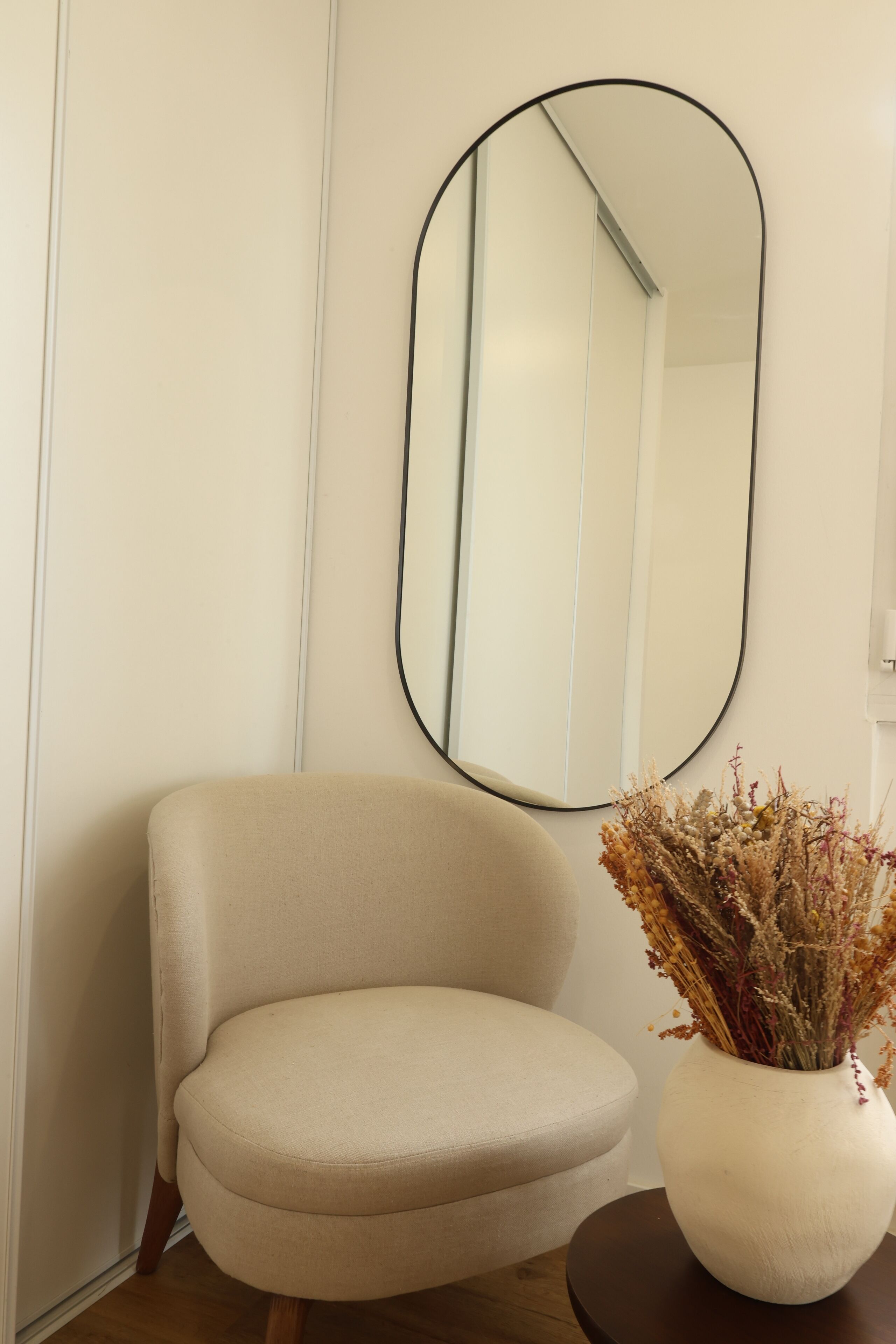 Een beige fauteuil, een ronde tafel met een vaas gedroogde bloemen, en een grote spiegel aan de muur.