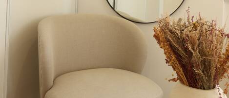 A beige armchair, a round table with a vase of dried flowers, and a large mirror on the wall.
