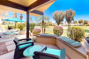 Relax on the patio with wicker chairs, a cushioned bench, and a table, surrounded by lush trees and shrubs.