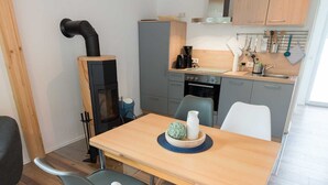 A compact kitchen with a wood stove, grey cabinets, and a dining area with a wooden table and two chairs. - Ferienhaus Harzblüte (Blankenburg (Harz))