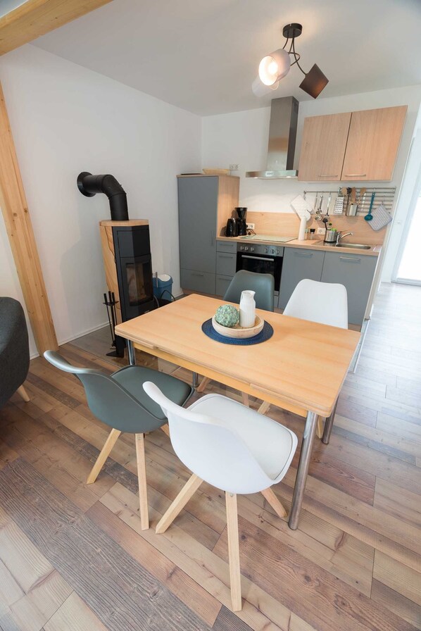 A compact kitchen with a wood stove, grey cabinets, and a dining area with a wooden table and two chairs. - Ferienhaus Harzblüte (Blankenburg (Harz))