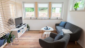 A modern living room with a grey sofa, a TV on a white stand, a small round table, and a window with blinds. - Ferienhaus Harzblüte (Blankenburg (Harz))