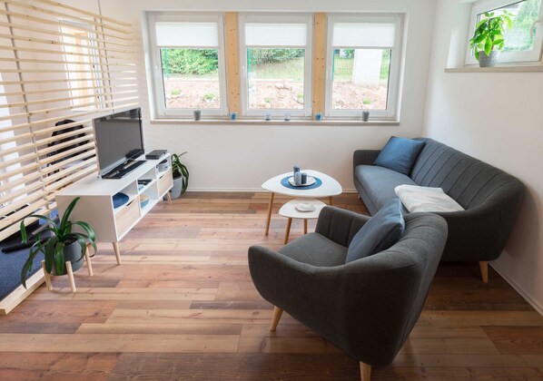 Un salon moderne avec un canapé gris, un téléviseur posé sur un meuble blanc, une petite table ronde et une fenêtre équipée de stores.