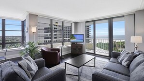 A modern living room with a sofa set, a coffee table, and a TV. Large windows offer a view of the city and a balcony. - Wing Beachfront Condo | Pool, Gym & Tennis Courts (Marco Island)