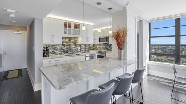 A modern kitchen with white cabinets, a granite countertop, and a large island with bar stools. - Wing Beachfront Condo | Pool, Gym & Tennis Courts (Marco Island)