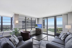 A modern living room with a sofa set, a coffee table, and a TV. Large windows offer a view of the city and a balcony.