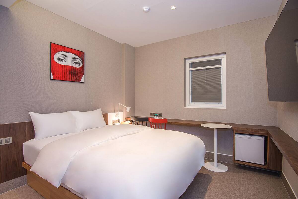Habitación de hotel con una cama grande, mesita de noche, lámpara y un pequeño escritorio con una silla.