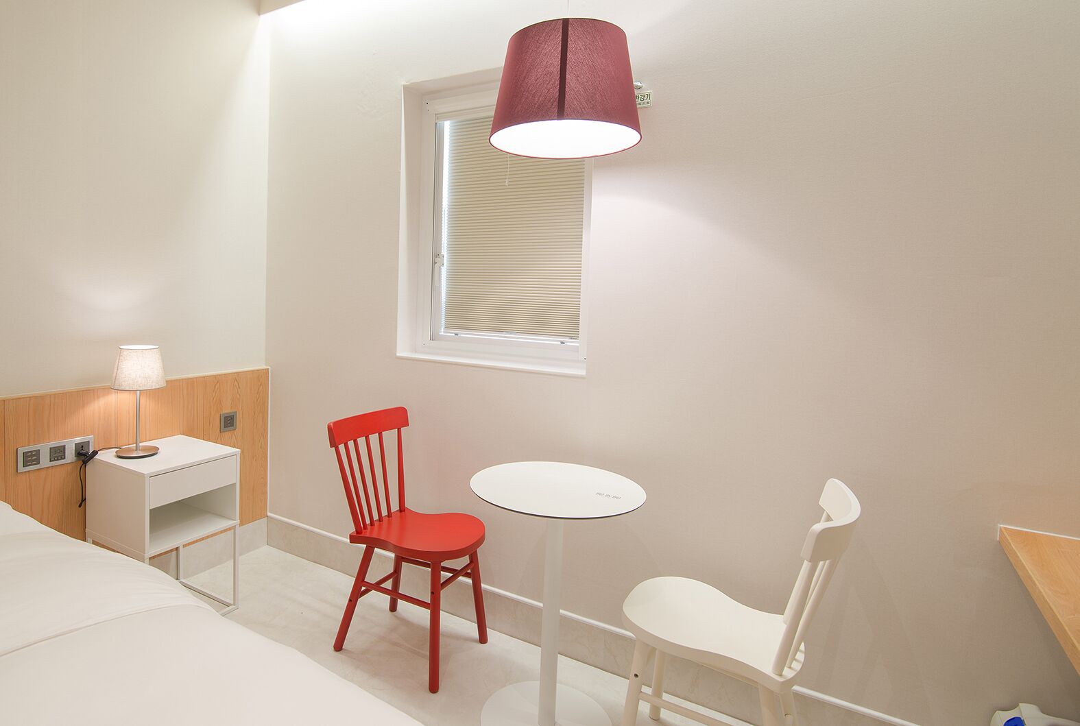 Habitación de hotel con una cama, una silla roja, una silla blanca, una mesita, una lámpara, una ventana con persianas y una lámpara de pared.