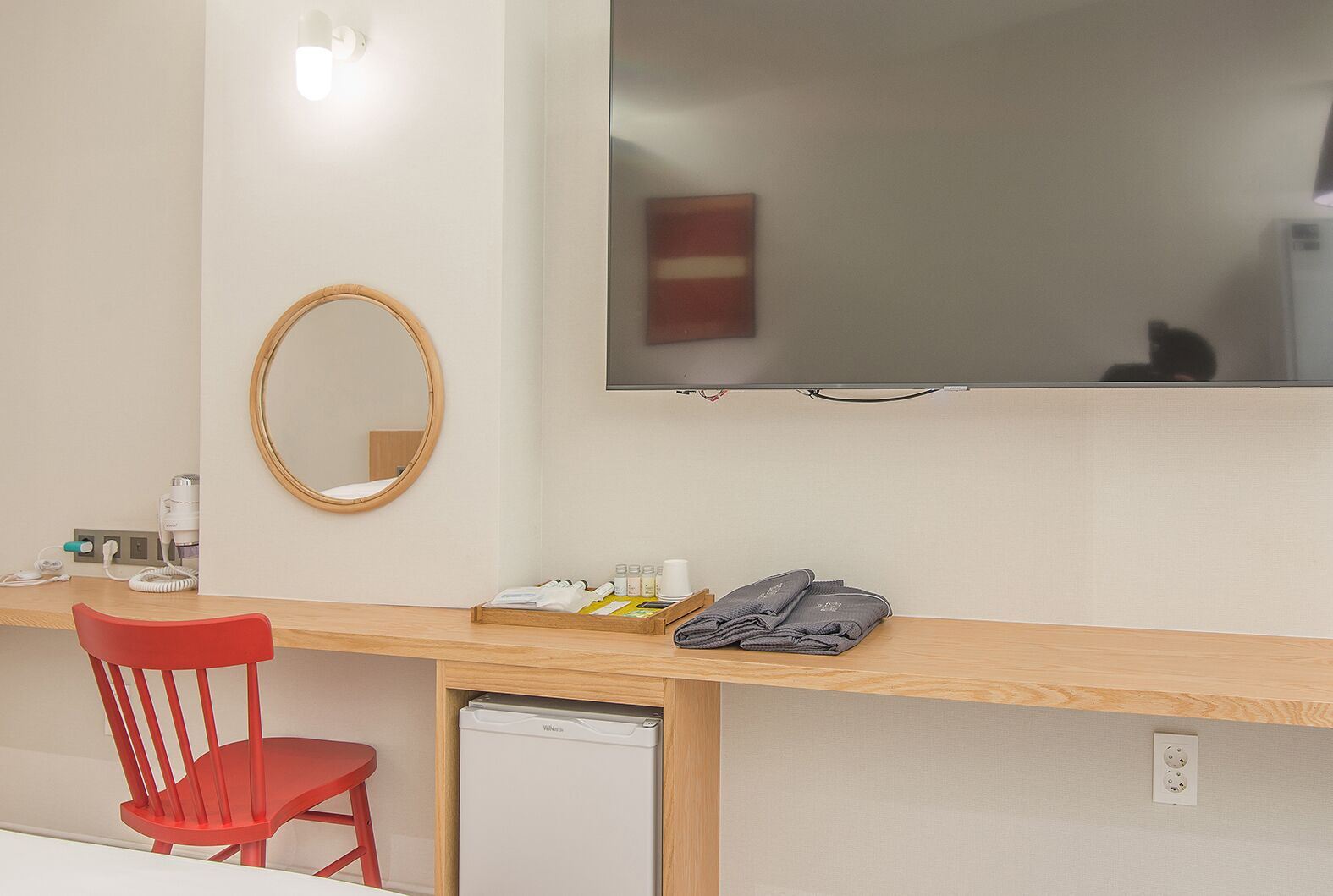 A hotel room with a wooden desk, a red chair, a small refrigerator, a mirror, and a flat-screen TV mounted on the wall.