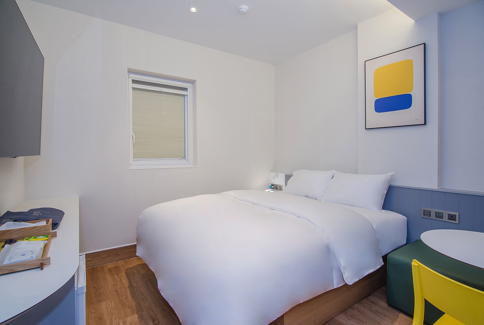 A modern hotel room with a large bed, a bedside table, a small desk, and a bathroom with a mirror and sink.