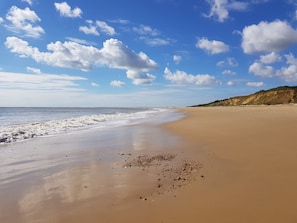 Una ampla platja de sorra daurada amb ones suaus i un cel blau clar amb algunes boires disperses.