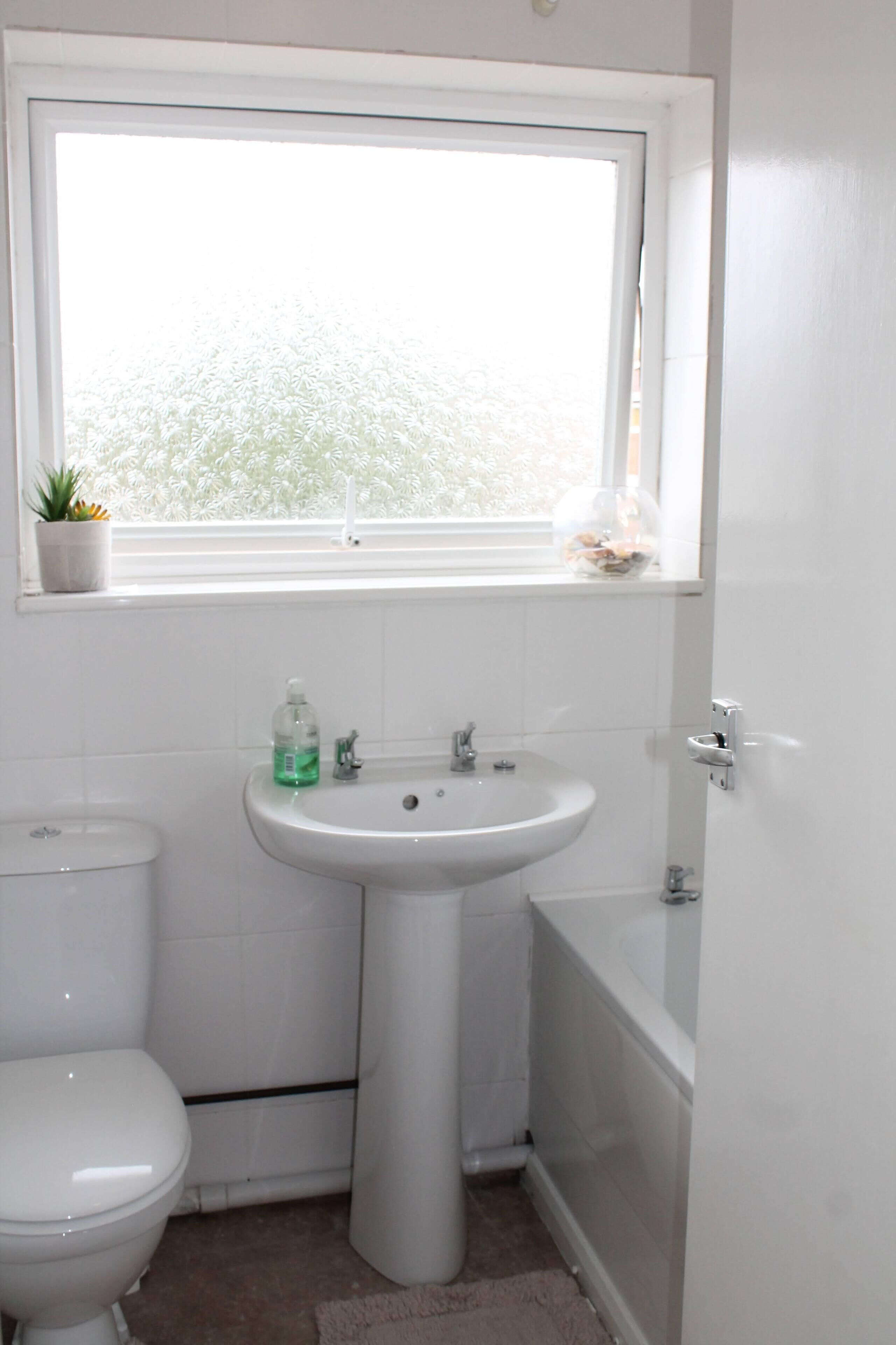 Un baño pequeño y blanco con inodoro, lavabo y bañera. Hay una ventana sobre el lavabo, y una planta pequeña en el alféizar.