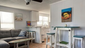 A living room with a grey sofa, a wooden coffee table, and a kitchen area with white cabinets and a blue-framed glass shelf.