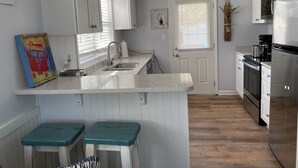 A kitchen with white cabinets, a stainless steel refrigerator, a stove, and a sink. There are two stools and a painting on the wall.