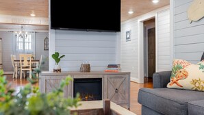 A living room with a fireplace, a flat-screen TV mounted above it, a grey sofa with a decorative pillow, and a dining area in the background.