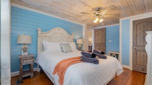 A bedroom with a wooden ceiling, a bed with white bedding, a ceiling fan, and a blue wall.