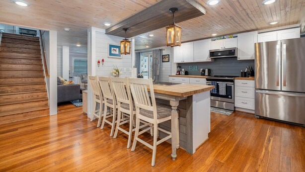 A modern kitchen with wooden flooring, a central island with chairs, stainless steel appliances, and pendant lighting.