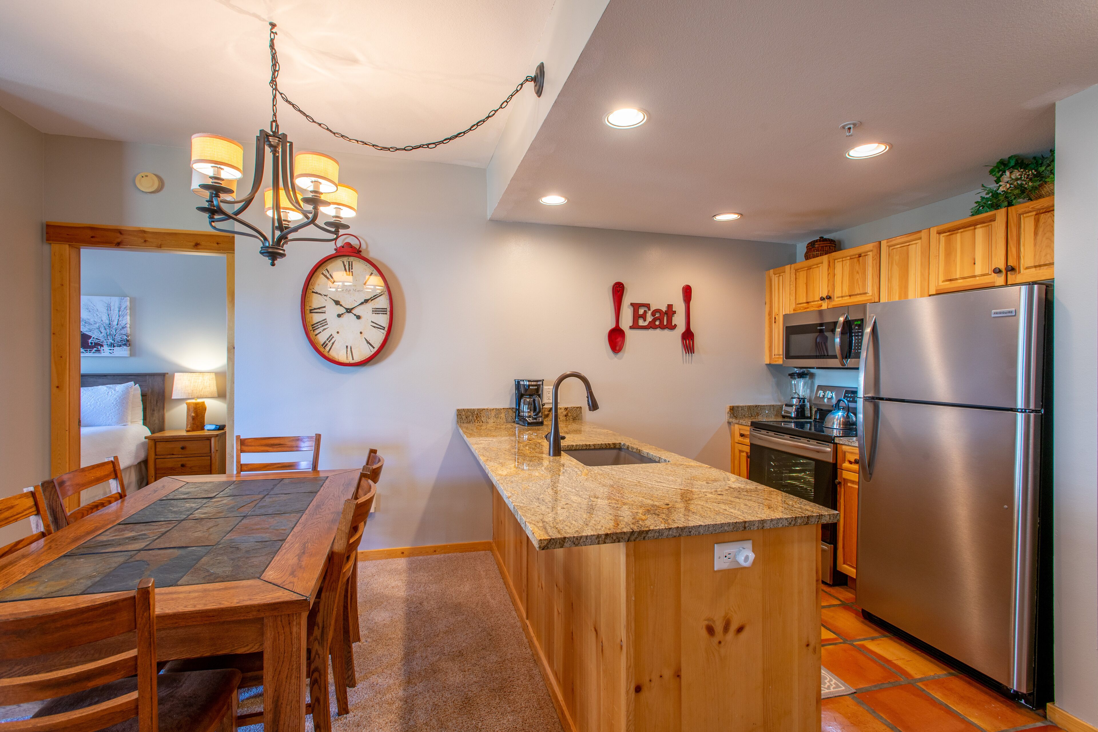 A kitchen with wooden cabinets, a granite countertop, a stainless steel refrigerator, and a clock on the wall.