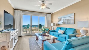 A spacious living room with a blue sofa, a coffee table, and a view of the beach through large sliding doors. - WHERE THE GULF MEETS YOUR LIVING ROOM! (Ft Walton Beach)