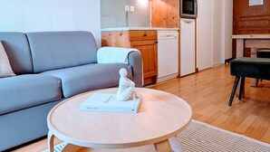 A modern living room with a grey sofa, a round wooden coffee table with a book and a figurine, and a kitchen area with wooden cabinets and a microwave.