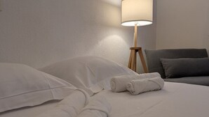 A neatly made bed with white linens and rolled towels, a grey sofa, and a bedside lamp.
